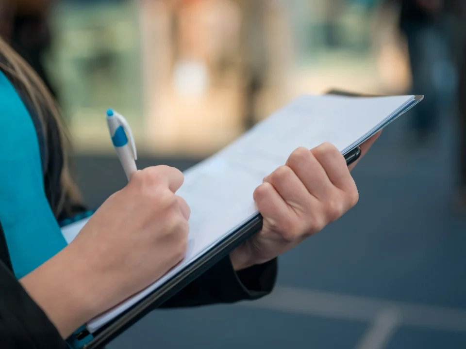 Person holding a pen and writing on a clipboard with papers, wearing a blue garment and dark jacket, with a blurred background