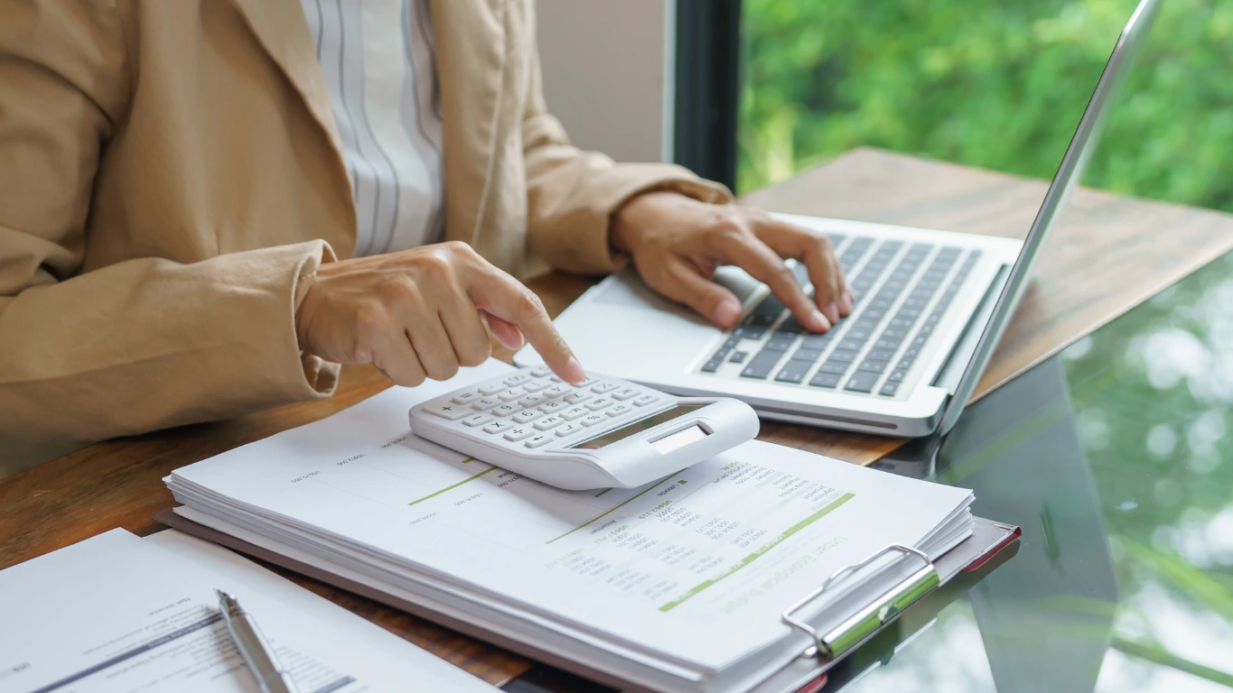 Close-up of a person using a calculator and laptop at a wooden desk, with documents and charts on a clipboard in front of them.